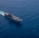 An aerial photograph of a large US Navy amphibious assault ship underway in open blue ocean waters. The flight deck is populated with multiple rotary-wing and fixed-wing aircraft, and the vessel is leaving a visible white wake behind it. A single aircraft appears to be airborne in the upper portion of the image.
