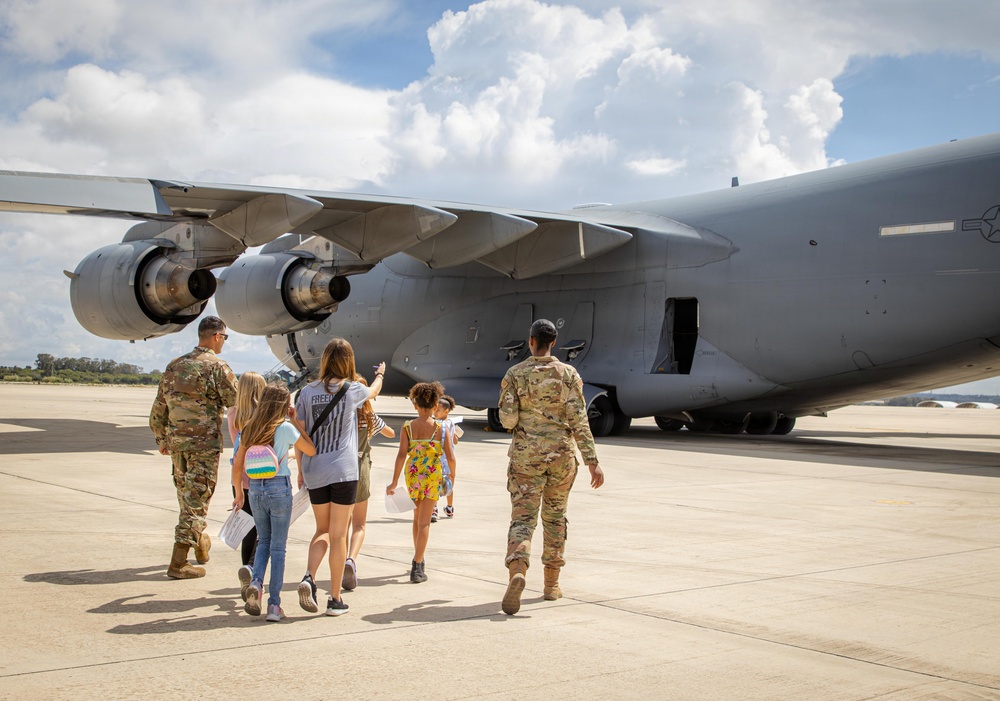 Two military personnel in OCP (Operational Camouflage Pattern) uniforms are escorting a group of civilians, including several children, toward a large gray military transport aircraft on a tarmac. The scene appears to be a family day, air show, or base open house event where families are being given a close-up tour of the aircraft. The setting is a sunny day with partial cloud cover at what appears to be a military airfield.