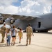 Two military personnel in OCP (Operational Camouflage Pattern) uniforms are escorting a group of civilians, including several children, toward a large gray military transport aircraft on a tarmac. The scene appears to be a family day, air show, or base open house event where families are being given a close-up tour of the aircraft. The setting is a sunny day with partial cloud cover at what appears to be a military airfield.