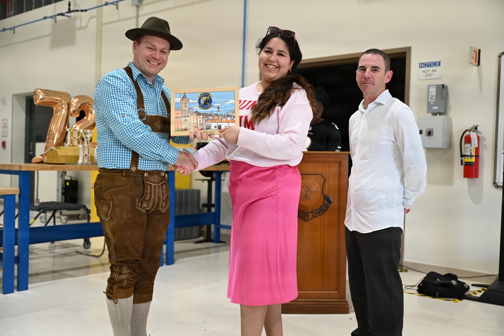 Three individuals are posed in what appears to be a military or institutional facility, with two of them shaking hands while holding a framed artwork or plaque. The man on the left is dressed in traditional Bavarian lederhosen attire, suggesting a cultural or themed event. A military unit emblem is visible on a wooden podium in the background.