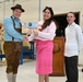 Three individuals are posed in what appears to be a military or institutional facility, with two of them shaking hands while holding a framed artwork or plaque. The man on the left is dressed in traditional Bavarian lederhosen attire, suggesting a cultural or themed event. A military unit emblem is visible on a wooden podium in the background.