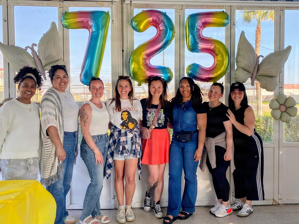 A group of eight women pose together in front of large rainbow foil number balloons reading '725' flanked by white butterfly and flower balloon decorations. The setting appears to be an indoor/covered outdoor space with large windows, suggesting a celebratory event such as a birthday party or milestone gathering. The group is smiling and appears to be in a festive, casual atmosphere.