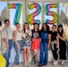 A group of eight women pose together in front of large rainbow foil number balloons reading '725' flanked by white butterfly and flower balloon decorations. The setting appears to be an indoor/covered outdoor space with large windows, suggesting a celebratory event such as a birthday party or milestone gathering. The group is smiling and appears to be in a festive, casual atmosphere.