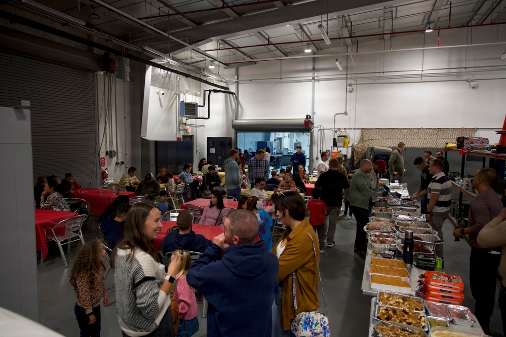 A large indoor gathering, likely a community or workplace potluck/holiday meal event, is taking place in what appears to be a warehouse or industrial facility. Multiple families including children are present, with long buffet-style tables on the right side loaded with food in aluminum trays. Dining tables covered with red tablecloths are set up on the left side where people are seated and eating.