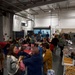 A large indoor gathering, likely a community or workplace potluck/holiday meal event, is taking place in what appears to be a warehouse or industrial facility. Multiple families including children are present, with long buffet-style tables on the right side loaded with food in aluminum trays. Dining tables covered with red tablecloths are set up on the left side where people are seated and eating.