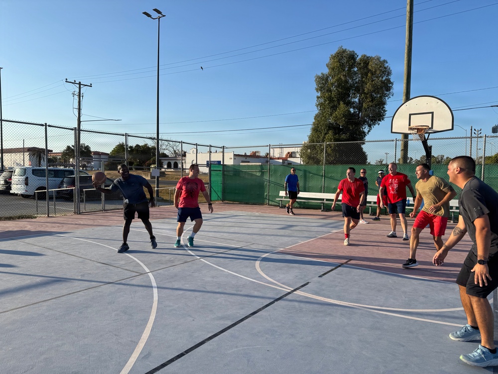 A group of men are playing an informal outdoor basketball game on a paved court during what appears to be late afternoon or early morning based on the low-angle sunlight. Players are divided into two apparent teams, with some wearing red shirts and others in darker or neutral-colored clothing. The court is enclosed by a chain-link fence and features standard basketball infrastructure including a visible backboard and hoop.