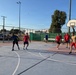 A group of men are playing an informal outdoor basketball game on a paved court during what appears to be late afternoon or early morning based on the low-angle sunlight. Players are divided into two apparent teams, with some wearing red shirts and others in darker or neutral-colored clothing. The court is enclosed by a chain-link fence and features standard basketball infrastructure including a visible backboard and hoop.