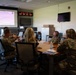 A group of military personnel in camouflage uniforms are gathered around a conference table for a briefing or meeting. A presentation titled 'DAF Spectrum of Resilience' is displayed on a large projection screen at the front of the room. The setting appears to be a military operations or conference room with multiple digital clocks on the wall.