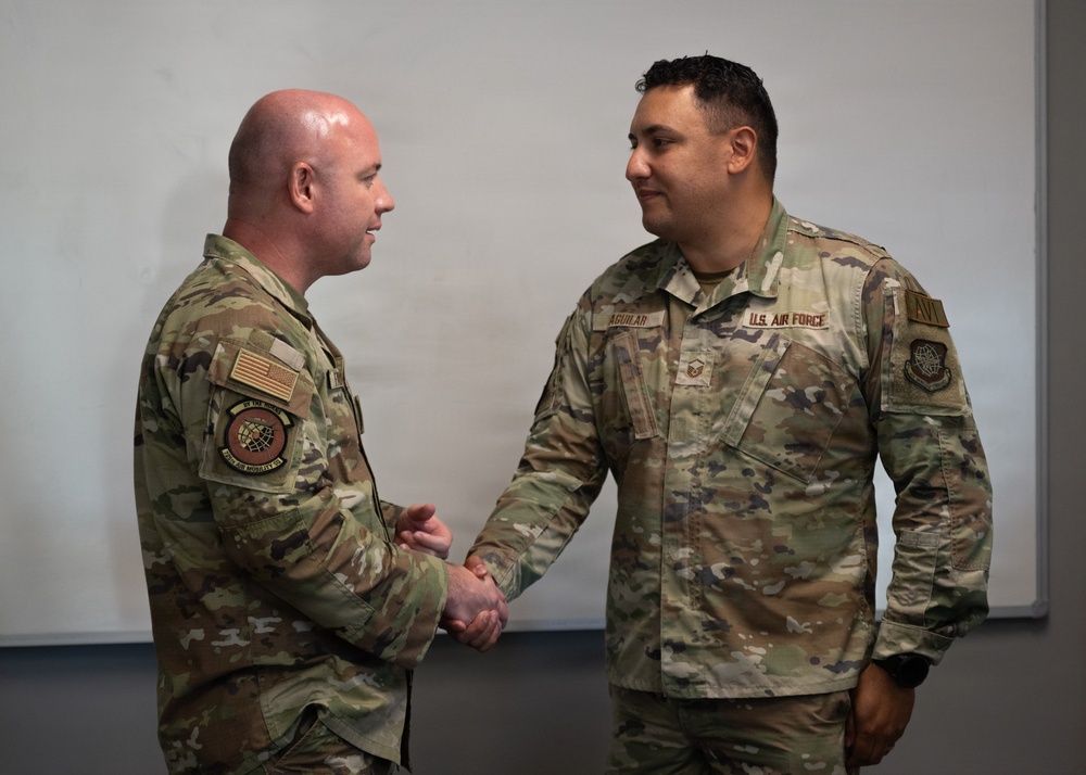 Two U.S. Air Force personnel in OCP (Operational Camouflage Pattern) uniforms are shaking hands in what appears to be an indoor military setting. The individual on the right has a visible name tape reading 'AGUILAR' and a 'U.S. AIR FORCE' tape, along with a Technical Sergeant rank insignia. Both personnel wear unit patches associated with Air Mobility Command.