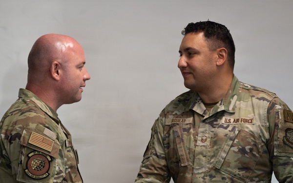 Two U.S. Air Force personnel in OCP (Operational Camouflage Pattern) uniforms are shaking hands in what appears to be an indoor military setting. The individual on the right has a visible name tape reading 'AGUILAR' and a 'U.S. AIR FORCE' tape, along with a Technical Sergeant rank insignia. Both personnel wear unit patches associated with Air Mobility Command.