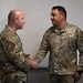 Two U.S. Air Force personnel in OCP (Operational Camouflage Pattern) uniforms are shaking hands in what appears to be an indoor military setting. The individual on the right has a visible name tape reading 'AGUILAR' and a 'U.S. AIR FORCE' tape, along with a Technical Sergeant rank insignia. Both personnel wear unit patches associated with Air Mobility Command.