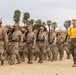 A formation of military recruits in desert camouflage (MARPAT) uniforms marches in disciplined rows while carrying rifles. A drill instructor wearing a distinctive yellow t-shirt and MARPAT trousers walks alongside the formation, mouth open as if issuing commands. The setting appears to be a military training depot, consistent with U.S. Marine Corps recruit training.