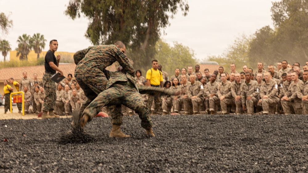 Two Marines in camouflage uniforms are engaged in a hand-to-hand combat demonstration on a dark gravel surface, while a large formation of uniformed Marines watches from the background. An instructor in a black shirt observes from the side, and several individuals in yellow shirts are also present among the spectators. The setting appears to be a military base, likely during a martial arts or combat training demonstration.