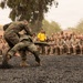 Two Marines in camouflage uniforms are engaged in a hand-to-hand combat demonstration on a dark gravel surface, while a large formation of uniformed Marines watches from the background. An instructor in a black shirt observes from the side, and several individuals in yellow shirts are also present among the spectators. The setting appears to be a military base, likely during a martial arts or combat training demonstration.