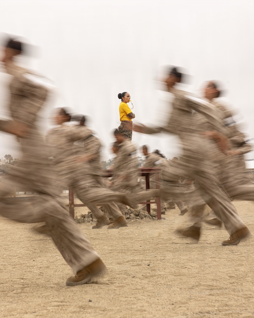 A motion-blur photograph captures a group of military recruits in tan/camouflage uniforms running past a stationary instructor who stands in sharp focus wearing a yellow t-shirt with arms crossed and a whistle in her mouth. The deliberate slow-shutter-speed technique renders the running recruits as blurred streaks while keeping the instructor pin-sharp, creating a strong visual contrast between movement and stillness. The setting appears to be a sandy outdoor training ground, consistent with a military boot camp environment.
