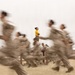 A motion-blur photograph captures a group of military recruits in tan/camouflage uniforms running past a stationary instructor who stands in sharp focus wearing a yellow t-shirt with arms crossed and a whistle in her mouth. The deliberate slow-shutter-speed technique renders the running recruits as blurred streaks while keeping the instructor pin-sharp, creating a strong visual contrast between movement and stillness. The setting appears to be a sandy outdoor training ground, consistent with a military boot camp environment.