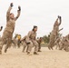 A large group of military personnel in desert camouflage uniforms are performing physical training exercises on a sandy drill field. The recruits, both male and female, are captured mid-jump during what appears to be a group calisthenics or physical fitness drill. An instructor in a yellow shirt is visible in the background overseeing the exercise.