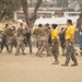 A military training exercise is taking place on a sandy outdoor field, likely at a U.S. Marine Corps base. Recruits or trainees in desert camouflage uniforms are engaged in hand-to-hand combat or martial arts drills using padded shields, while instructors in yellow shirts supervise the activity. The background shows trees, vehicles, and base buildings consistent with a Southern California military installation.