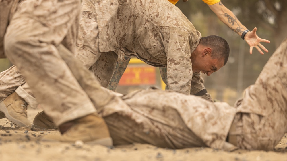 The image shows military personnel in desert camouflage (MARPAT) uniforms engaged in a ground combat or martial arts training exercise on a sandy surface. One individual is positioned over another who is on the ground, suggesting a combatives or hand-to-hand combat drill. A third person in a yellow shirt is partially visible in the background, likely serving as an instructor or observer.