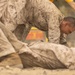 The image shows military personnel in desert camouflage (MARPAT) uniforms engaged in a ground combat or martial arts training exercise on a sandy surface. One individual is positioned over another who is on the ground, suggesting a combatives or hand-to-hand combat drill. A third person in a yellow shirt is partially visible in the background, likely serving as an instructor or observer.
