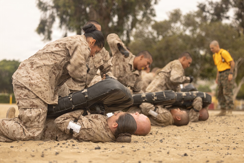 The image shows a group of military personnel in MARPAT (Marine pattern) camouflage uniforms engaged in what appears to be a combat training or martial arts exercise on a sandy outdoor surface. Multiple pairs of trainees are practicing ground-based combative techniques, with one person kneeling over another who is lying face-up on the ground. A supervisor in a yellow shirt observes the exercise in the background.