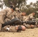 The image shows a group of military personnel in MARPAT (Marine pattern) camouflage uniforms engaged in what appears to be a combat training or martial arts exercise on a sandy outdoor surface. Multiple pairs of trainees are practicing ground-based combative techniques, with one person kneeling over another who is lying face-up on the ground. A supervisor in a yellow shirt observes the exercise in the background.