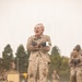 A group of military personnel in desert camouflage uniforms (MARPAT pattern) are engaged in a physical training exercise on a sandy field. The foreground subject, a young male with a shaved head, is shouting with arms crossed over his chest while appearing to carry or be carried by another person behind him. Multiple other service members are visible performing similar activities in the background.
