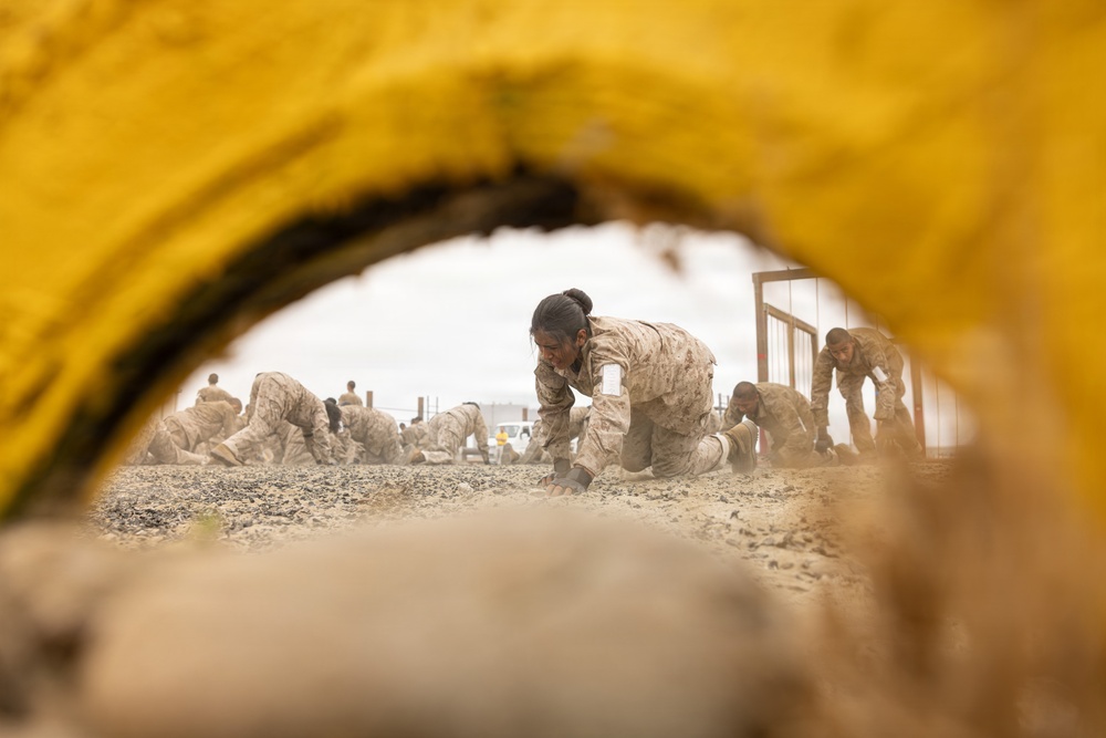 The image is photographed through a yellow circular tunnel obstacle, framing a group of military personnel in desert camouflage uniforms crawling on hands and knees across a sandy, gravelly terrain. The recruits or trainees appear to be participating in a physical training or obstacle course exercise. The foreground yellow tunnel element creates a natural frame, adding depth to the composition.