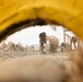 The image is photographed through a yellow circular tunnel obstacle, framing a group of military personnel in desert camouflage uniforms crawling on hands and knees across a sandy, gravelly terrain. The recruits or trainees appear to be participating in a physical training or obstacle course exercise. The foreground yellow tunnel element creates a natural frame, adding depth to the composition.