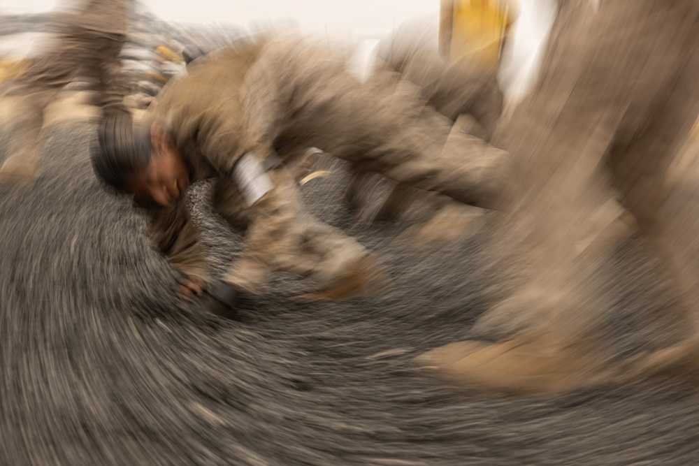 A heavily motion-blurred photograph captures what appears to be a person in earth-toned clothing surrounded by animals, likely during a dynamic or fast-moving event. The radial blur effect creates a swirling vortex pattern centered on the figure, suggesting rapid rotational camera movement or a long exposure technique. The image appears to document an interaction between a human and large animals, possibly in a herding or wrestling context.