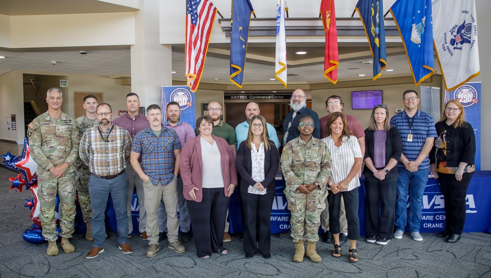 A group of approximately 16 individuals poses for a formal group photo in the lobby of the Kratos Conference Center at Naval Surface Warfare Center Crane (NSWC Crane). The group includes both military personnel in Army Combat Uniforms and civilians in business casual attire. Multiple military branch flags are displayed overhead, and NSWC Crane branded banners are visible in the background.