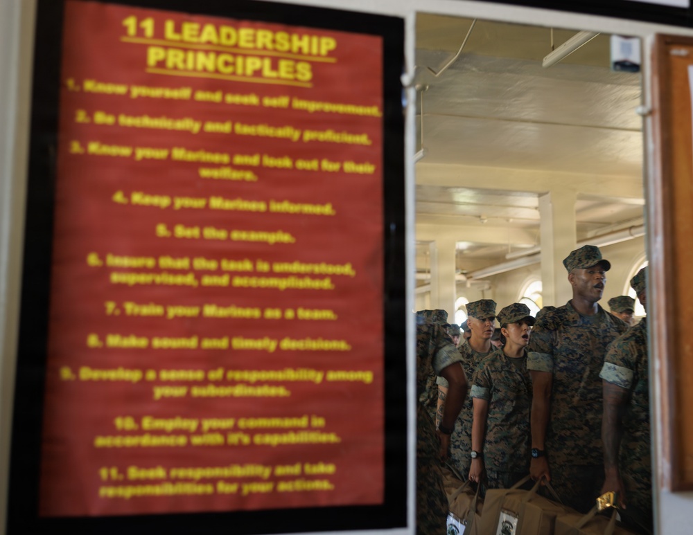 The image shows a red poster listing '11 Leadership Principles' in yellow text mounted on a wall, with a group of uniformed Marines visible through a doorway or window in the background. The Marines are wearing woodland MARPAT camouflage uniforms and covers (caps), and appear to be assembled in formation inside a building. The composition juxtaposes the leadership doctrine displayed on the poster with the military personnel it governs.