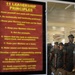 The image shows a red poster listing '11 Leadership Principles' in yellow text mounted on a wall, with a group of uniformed Marines visible through a doorway or window in the background. The Marines are wearing woodland MARPAT camouflage uniforms and covers (caps), and appear to be assembled in formation inside a building. The composition juxtaposes the leadership doctrine displayed on the poster with the military personnel it governs.
