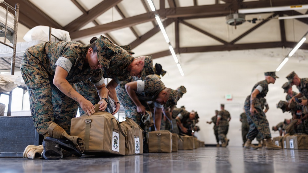 A group of military personnel in MARPAT (Marine digital camouflage) uniforms are bent over inspecting or packing tan/coyote-colored duffel bags arranged in a row on a polished floor inside a large indoor facility. The scene appears to depict a gear inspection, pre-deployment equipment check, or unit readiness drill. Multiple service members extend into the background, suggesting a large-scale organized formation activity.