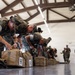 A group of military personnel in MARPAT (Marine digital camouflage) uniforms are bent over inspecting or packing tan/coyote-colored duffel bags arranged in a row on a polished floor inside a large indoor facility. The scene appears to depict a gear inspection, pre-deployment equipment check, or unit readiness drill. Multiple service members extend into the background, suggesting a large-scale organized formation activity.