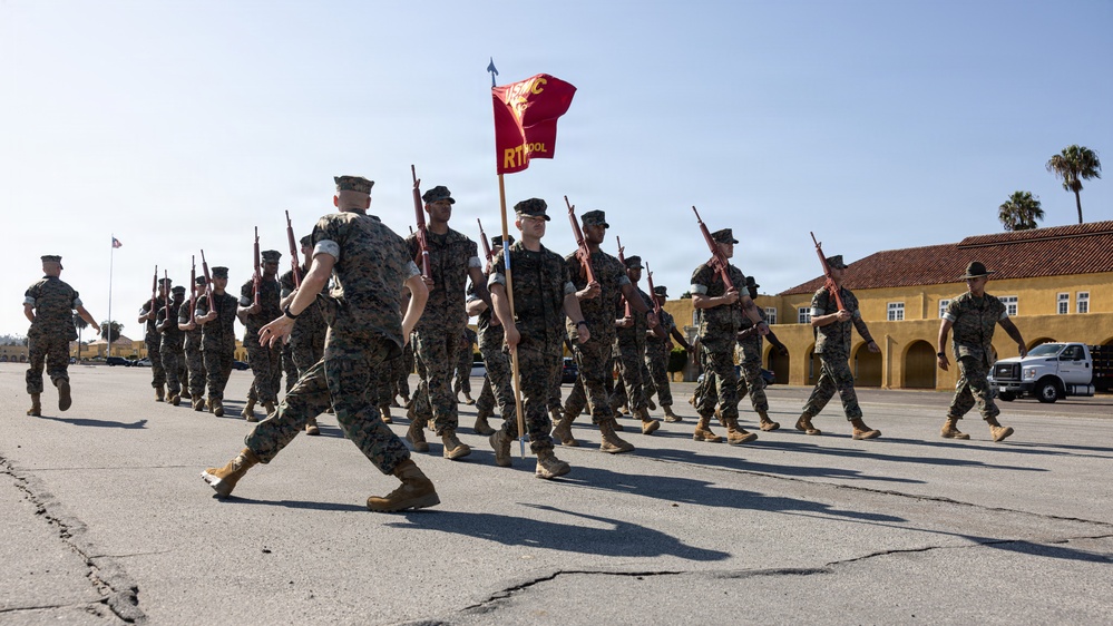 A formation of U.S. Marines in woodland MARPAT camouflage uniforms marches across a parade deck while carrying rifles at port arms. A red guidon flag bearing 'USMC' and 'R1' markings is carried at the front of the formation. The setting appears to be a Marine Corps recruit depot or training base, identifiable by the Spanish Colonial Revival-style architecture in the background.