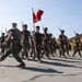 A formation of U.S. Marines in woodland MARPAT camouflage uniforms marches across a parade deck while carrying rifles at port arms. A red guidon flag bearing 'USMC' and 'R1' markings is carried at the front of the formation. The setting appears to be a Marine Corps recruit depot or training base, identifiable by the Spanish Colonial Revival-style architecture in the background.