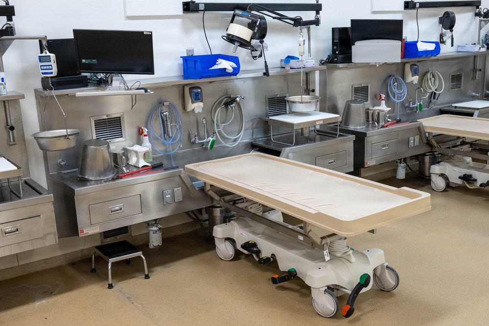 The image depicts a modern autopsy or mortuary suite with multiple workstations equipped with stainless steel surfaces, medical instruments, and computer monitors. A wheeled examination/transfer table with a beige plastic surface is positioned in the foreground. The facility appears clean, well-organized, and currently unoccupied.