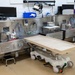 The image depicts a modern autopsy or mortuary suite with multiple workstations equipped with stainless steel surfaces, medical instruments, and computer monitors. A wheeled examination/transfer table with a beige plastic surface is positioned in the foreground. The facility appears clean, well-organized, and currently unoccupied.