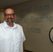 A middle-aged bald man with glasses and a short beard is posing and smiling in front of a wall sign reading 'Armed Forces Medical Examiner System.' He is wearing a white short-sleeve button-up shirt with a small dot pattern. An official seal/emblem is visible on the wall to his right.