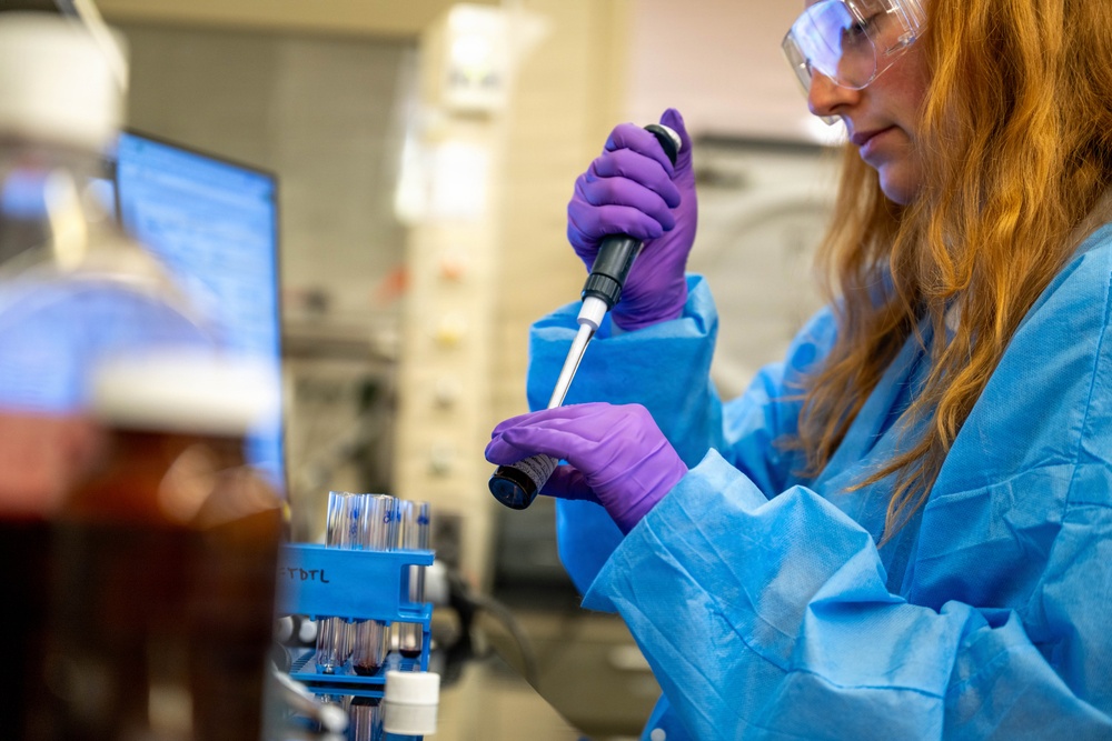 A female laboratory technician or researcher is working in a professional laboratory setting. She is using a micropipette to transfer liquid into or from a small vial or tube. She is wearing appropriate personal protective equipment including a blue lab coat, purple nitrile gloves, and safety goggles.