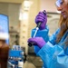 A female laboratory technician or researcher is working in a professional laboratory setting. She is using a micropipette to transfer liquid into or from a small vial or tube. She is wearing appropriate personal protective equipment including a blue lab coat, purple nitrile gloves, and safety goggles.