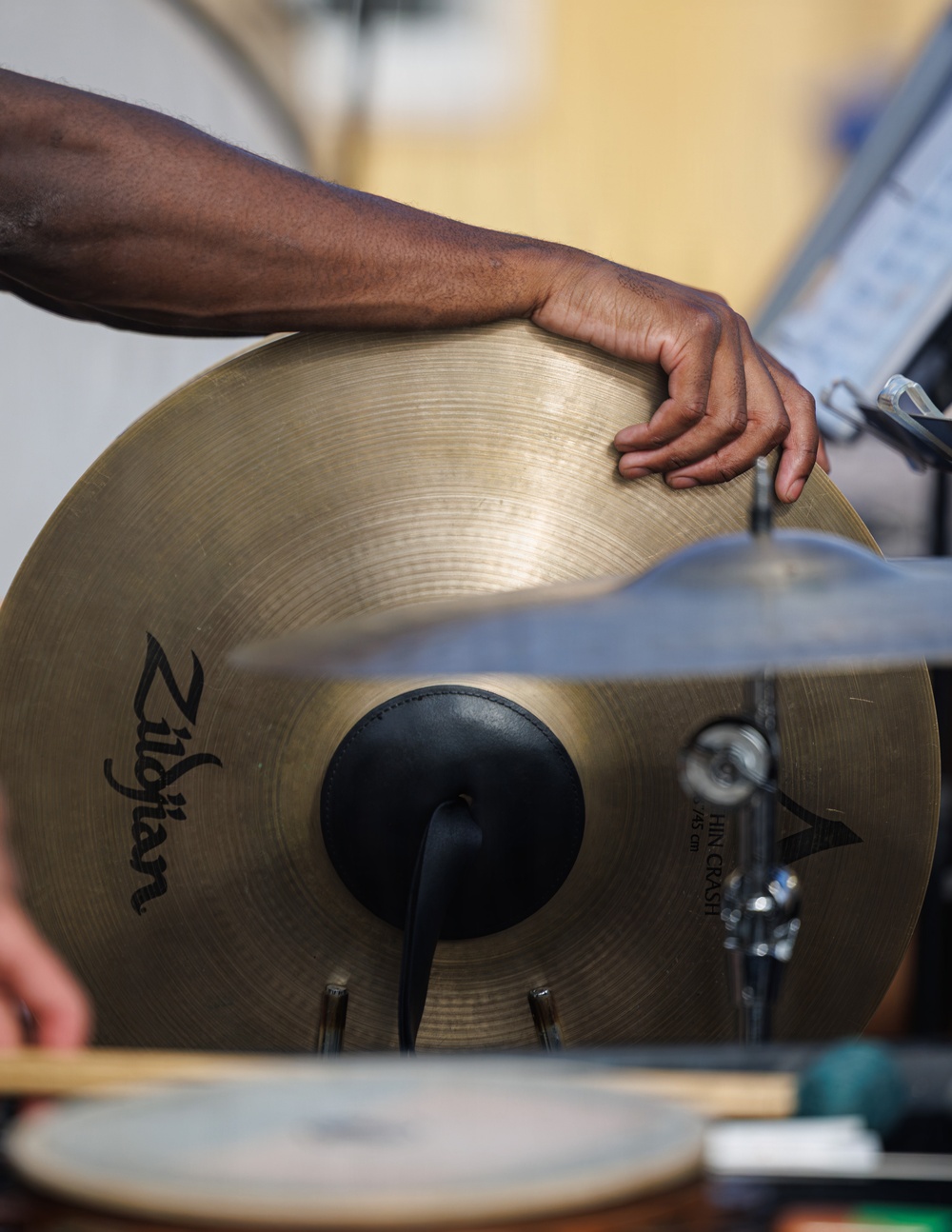 A close-up photograph of a drummer's hand resting on a Zildjian cymbal, likely muting or controlling its resonance after a strike. The image prominently features a large brass Zildjian cymbal with visible branding and a second cymbal partially visible in the foreground. A music stand with sheet music is partially visible in the blurred background.