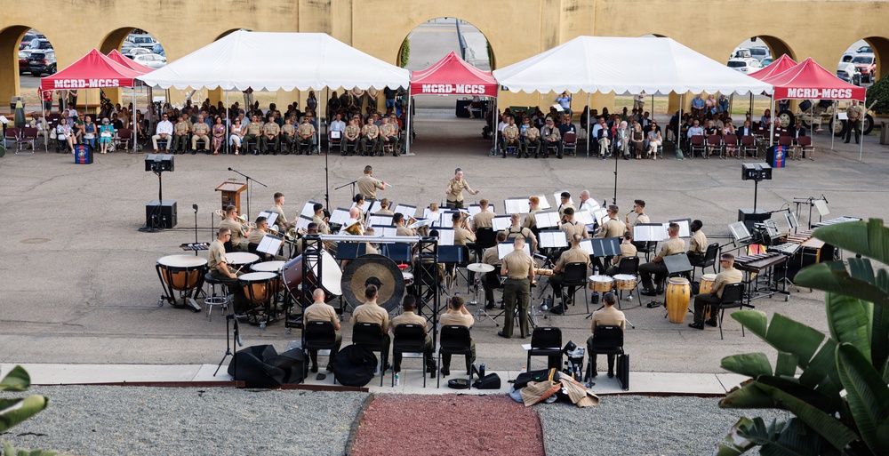 A military band, likely the MCRD (Marine Corps Recruit Depot) band, performs an outdoor concert in a courtyard setting. Musicians in tan/khaki uniforms are arranged in a semicircle with a conductor leading them, while an audience seated under red and white MCRD-MCCS branded canopy tents observes the performance. The event appears to be a formal military ceremony or concert held at a base facility.