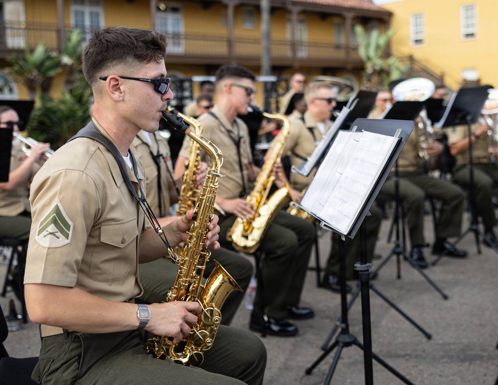 A U.S. Marine Corps band is performing outdoors in what appears to be a public square or plaza. The foreground features a Marine corporal playing an alto saxophone, with numerous other band members visible behind him playing various brass and woodwind instruments. The setting appears to be in a warm-climate location with colonial-style architecture visible in the background.