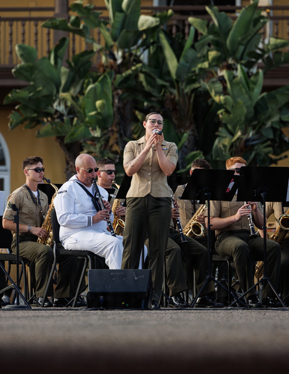 A military band performs an outdoor concert, with a standing vocalist in tan/khaki uniform singing into a microphone as the focal point. Musicians seated behind her play saxophones and clarinets, wearing a mix of tan military uniforms and one white Navy dress uniform. The performance takes place in front of a yellow building with tropical foliage in the background.