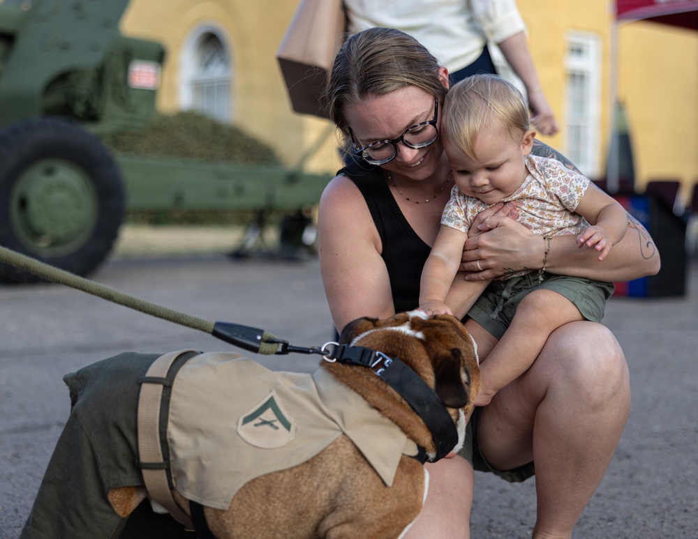 A woman wearing glasses holds a young toddler while both interact with a dog dressed in a military-style vest with a rank insignia patch. The scene appears to take place at an outdoor military event or base, with green military equipment visible in the background. The mood is warm and candid, capturing a lighthearted moment between the woman, child, and costumed dog.