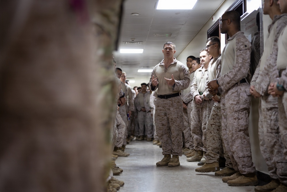 A military leader, dressed in a tan combat shirt and desert camouflage trousers, addresses a group of uniformed personnel standing in two lines along a corridor. The personnel are wearing MARPAT (Marine pattern) desert camouflage uniforms consistent with U.S. Marine Corps dress. The setting appears to be an indoor military facility, with the speaker gesturing as if delivering a briefing or motivational address.