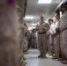 A military leader, dressed in a tan combat shirt and desert camouflage trousers, addresses a group of uniformed personnel standing in two lines along a corridor. The personnel are wearing MARPAT (Marine pattern) desert camouflage uniforms consistent with U.S. Marine Corps dress. The setting appears to be an indoor military facility, with the speaker gesturing as if delivering a briefing or motivational address.