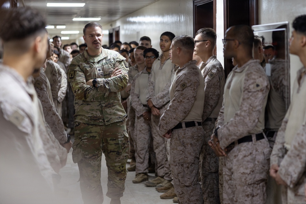 A U.S. Army officer in Operational Camouflage Pattern (OCP) uniform stands with arms crossed, appearing to address or brief a large group of military personnel in a hallway. The assembled group includes personnel wearing both OCP and Marine Corps MARPAT desert camouflage uniforms, suggesting a joint-service gathering. The setting appears to be an indoor military facility corridor.